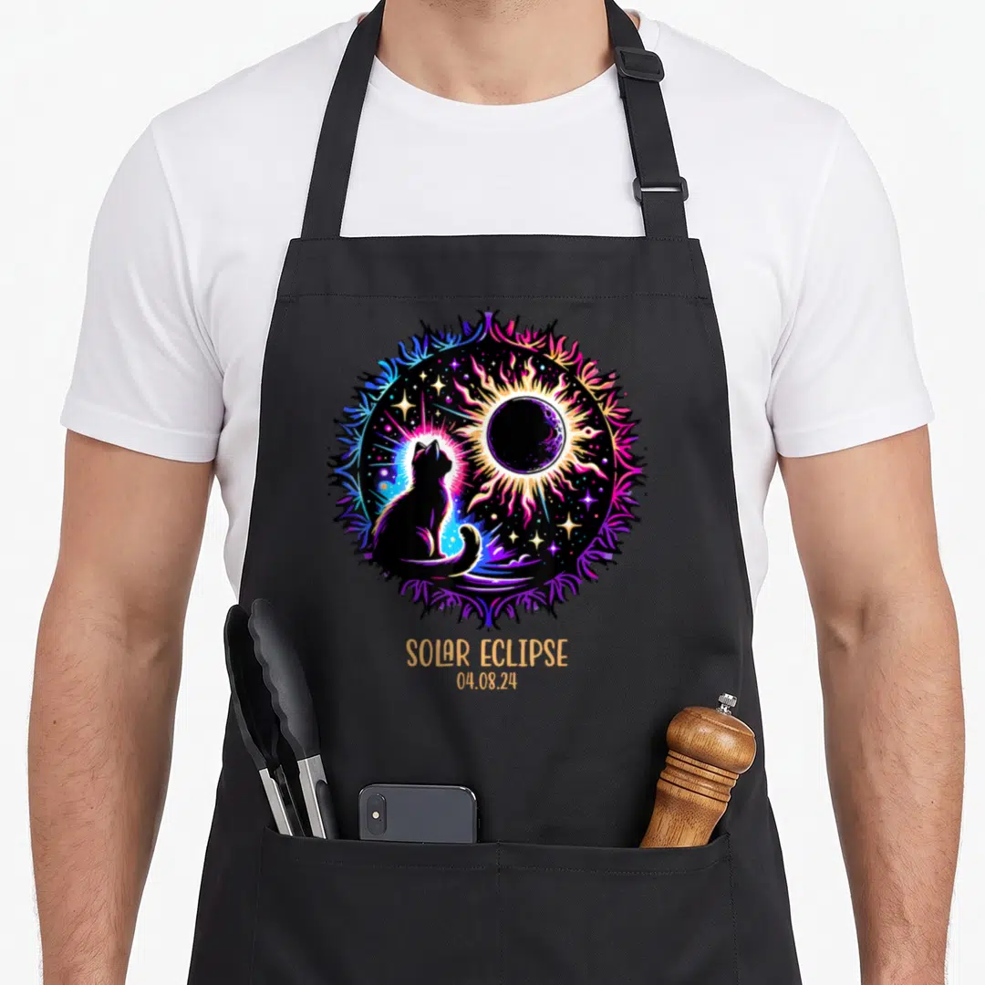View cat apron - Cat silhouette and solar eclipse in purple blue orange pink above SOLAR ECLIPSE 04.08.24 text — shown on a black cotton canvas apron against a white background