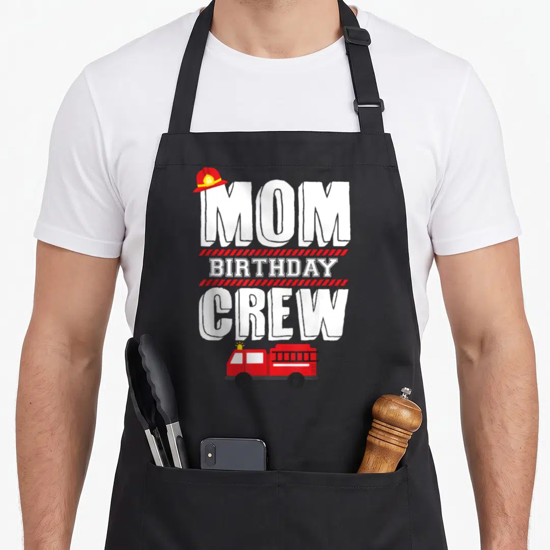 Fireman crew aprons - MOM BIRTHDAY CREW text with fire truck and helmet on grey background — shown on a black cotton canvas apron against a white background