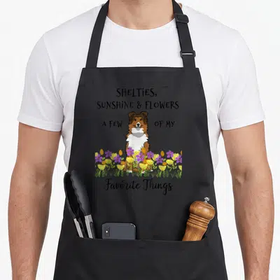 Shelty shetland sheepdog apron - Cartoon Sheltie sitting in purple yellow and green flowers with black text — shown on a black cotton canvas apron against a white background