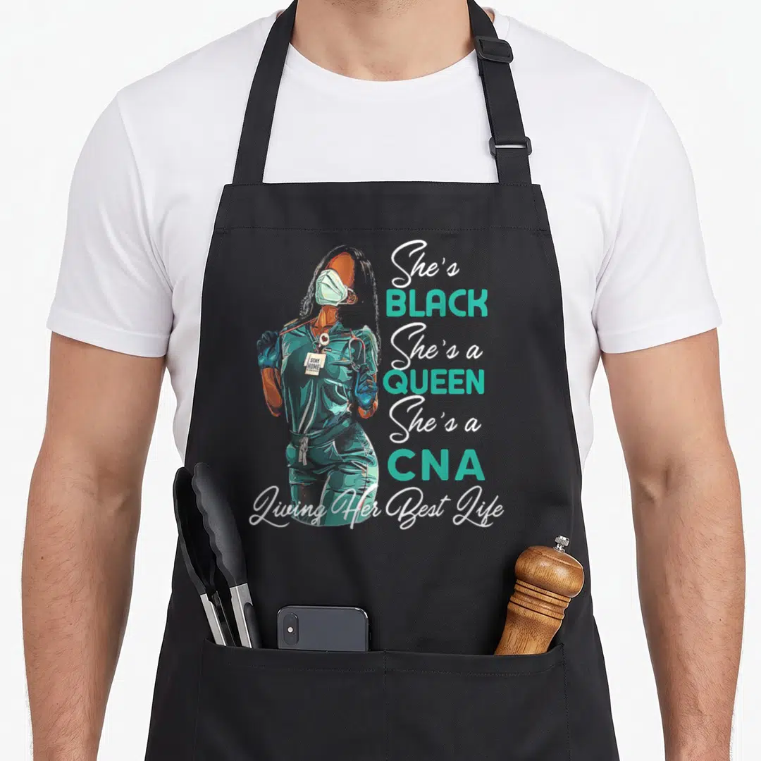 Cna queen apron - Cartoon woman in teal scrubs with stacked text She's Black She's a Queen She's a CNA — shown on a black cotton canvas apron against a white background