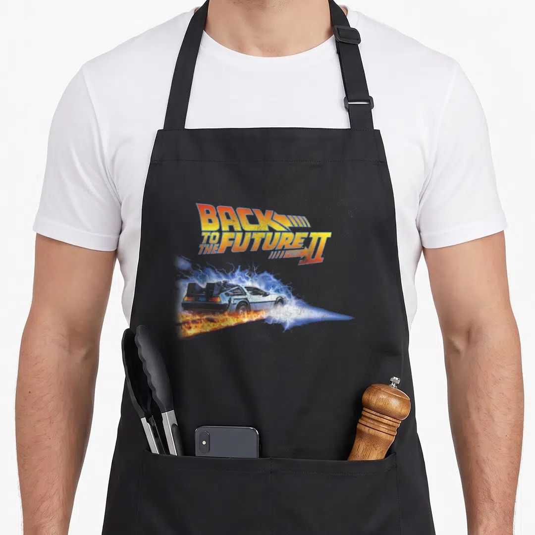 Flaming us aprons - Back To The Future II Part text above a DeLorean with fire trails and blue sparks — shown on a black cotton canvas apron against a white background