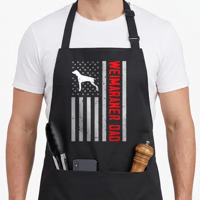 Weimaraner american flag apron - White dog silhouette on stars with red vertical WEIMARANER DAD text on stripes — shown on a black cotton canvas apron against a white background