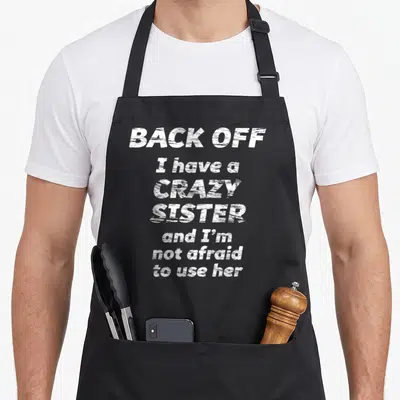 Funny sister use of apron - White distressed text reading Back Off I Have a Crazy Sister on gray background — shown on a black cotton canvas apron against a white background