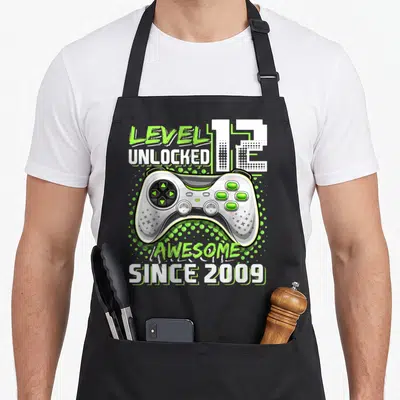 12th birthday video game apron - Game controller with LEVEL 12 UNLOCKED AWESOME SINCE 2009 text and lime green dots — shown on a black cotton canvas apron against a white background