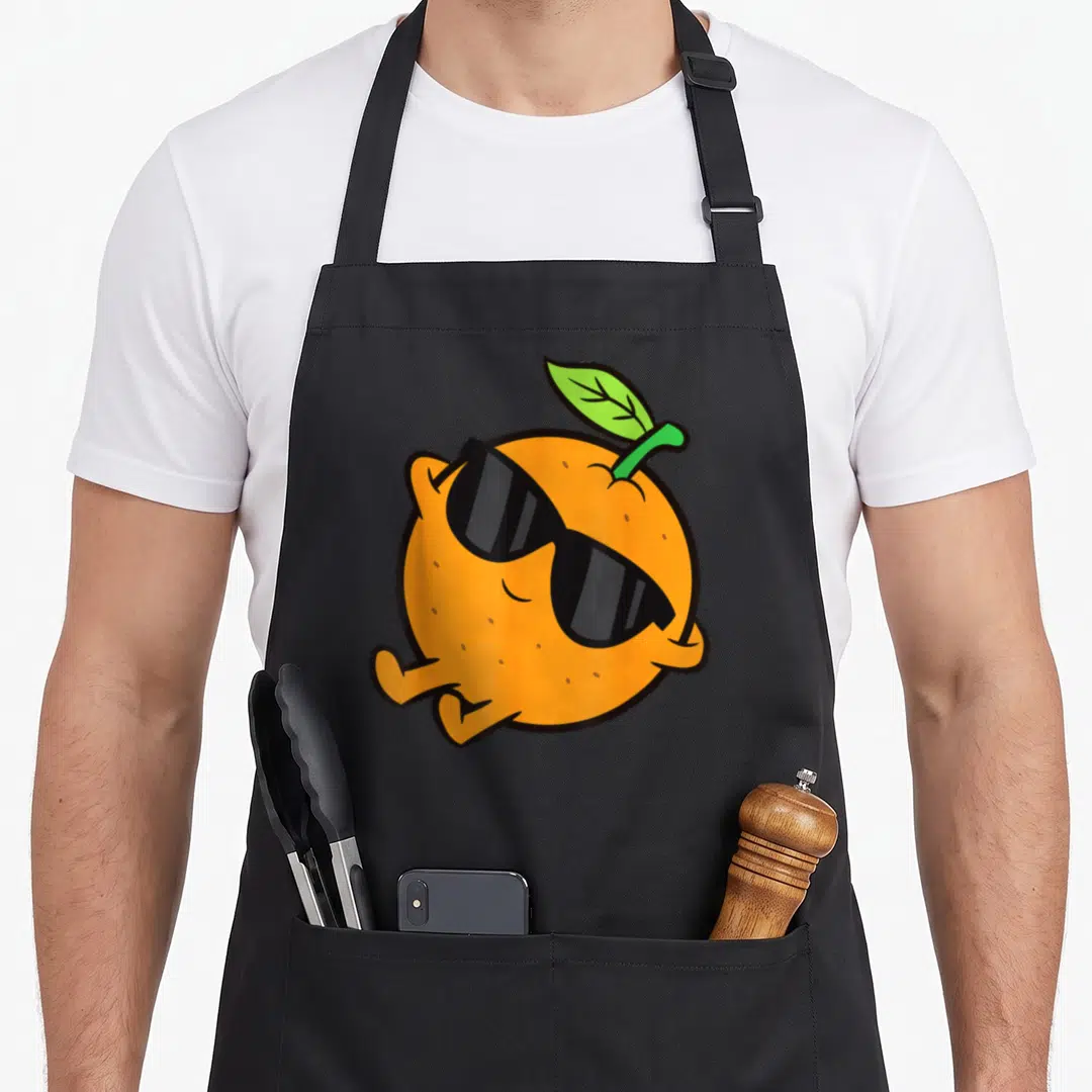Relaxing fruit apron - Cartoon orange character with black sunglasses, green leaf, and limbs in a relaxing pose — shown on a black cotton canvas apron against a white background