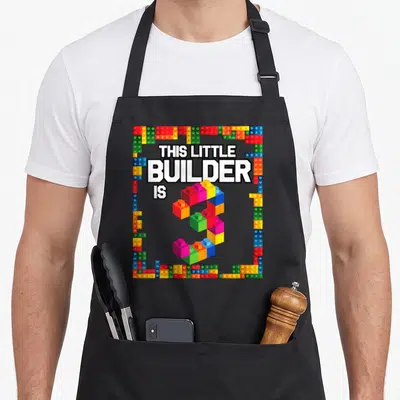 3rd building aprons - Number 3 made of red blue yellow green and white toy bricks with builder text — shown on a black cotton canvas apron against a white background