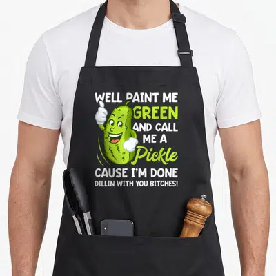 Paint pickle apron - Cartoon pickle thumbs up with well paint me green and call me a pickle text — shown on a black cotton canvas apron against a white background