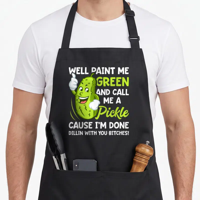 Paint pickle apron - Cartoon pickle thumbs up with well paint me green and call me a pickle text — shown on a black cotton canvas apron against a white background