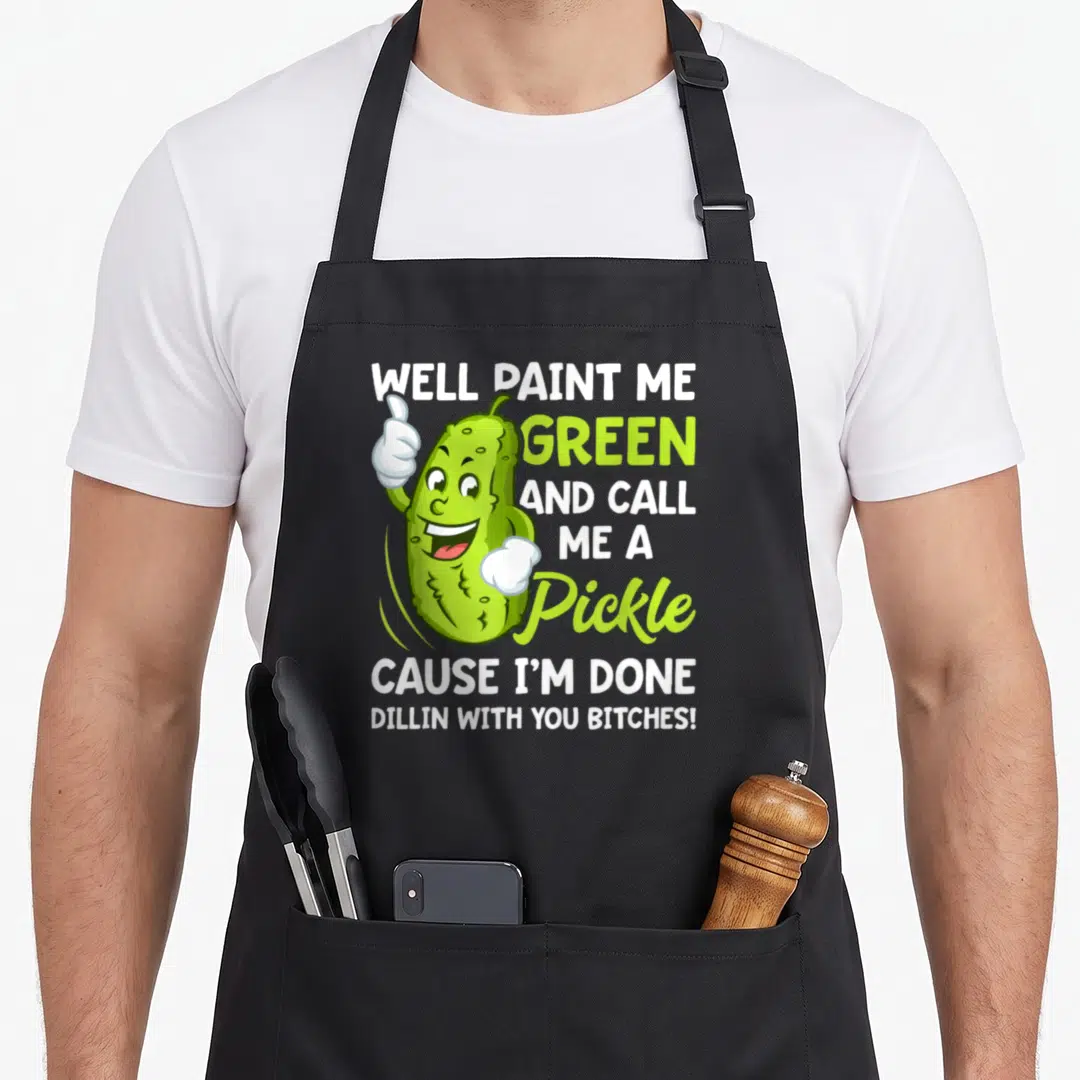 Paint pickle apron - Cartoon pickle thumbs up with well paint me green and call me a pickle text — shown on a black cotton canvas apron against a white background