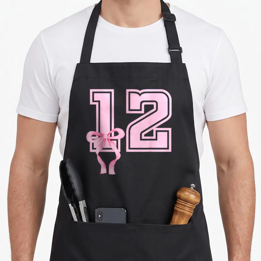 Funny birthday bow apron - Pink number 12 with watercolor coquette bow overlapping the bottom left corner — shown on a black cotton canvas apron against a white background
