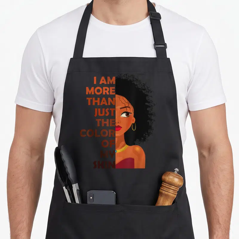 Skin novelty apron - Stacked orange and brown text next to illustrated woman with afro and gold hoop earrings — shown on a black cotton canvas apron against a white background