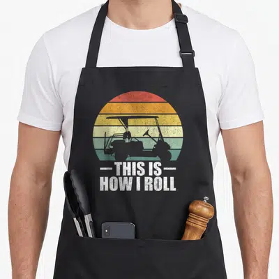 Roll golfer golf apron - Golf cart silhouette over red orange yellow green striped sun with white text THIS IS HOW I — shown on a black cotton canvas apron against a white background