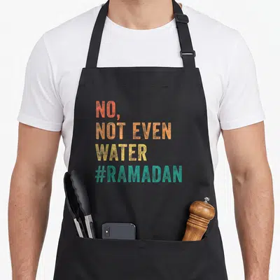 Mubarak ramadan apron - Stacked text reading No Not Even Water hashtag Ramadan in orange, gold and teal — shown on a black cotton canvas apron against a white background