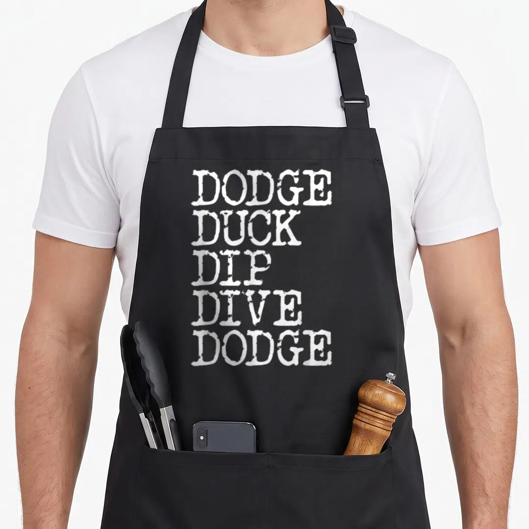 Dodge duck apron - White stacked text reading Dodge Duck Dip Dive Dodge — shown on a black cotton canvas apron against a white background