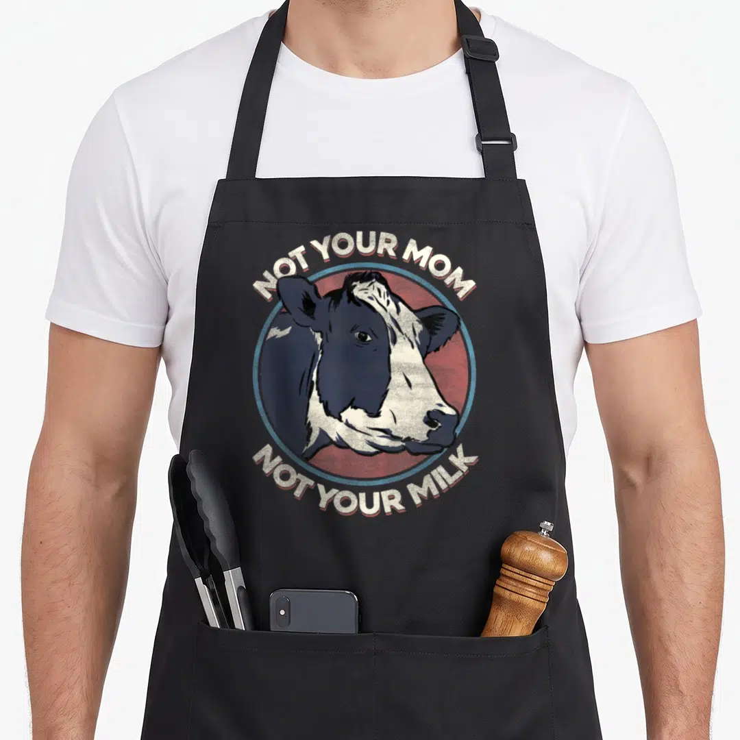 Milk vegan apron - Distressed cow head in red blue circle with text NOT YOUR MOM NOT YOUR MILK — shown on a black cotton canvas apron against a white background
