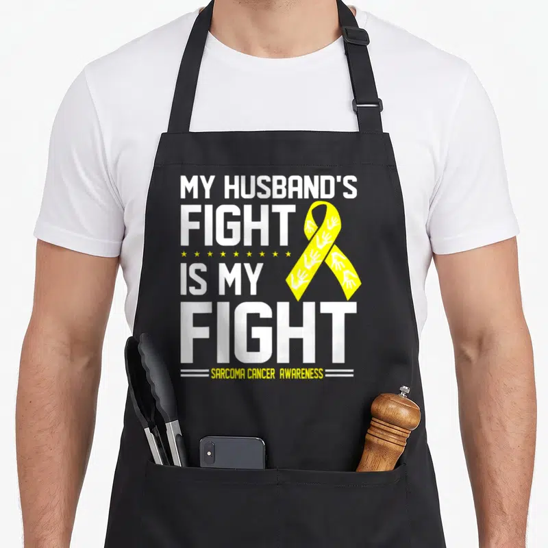 Sarcoma husband apron - White text reading MY HUSBAND'S FIGHT IS MY FIGHT next to a yellow ribbon with handprints — shown on a black cotton canvas apron against a white background