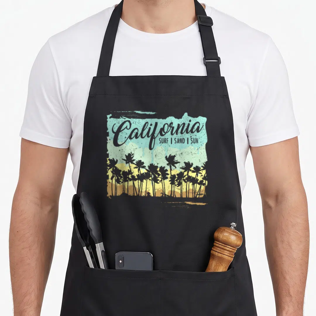 California summer aprons - Distressed grey blue yellow and brown graphic with palm trees and California text — shown on a black cotton canvas apron against a white background
