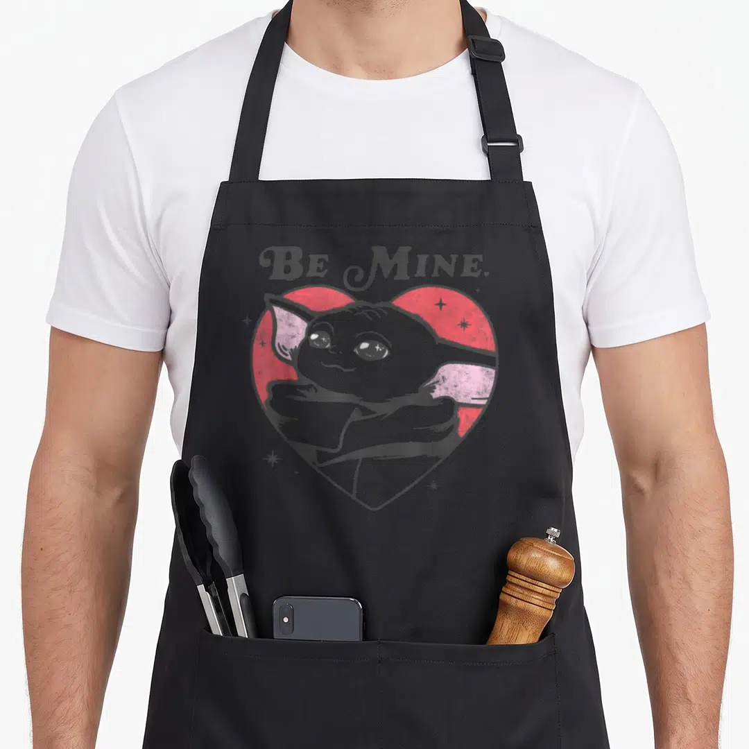 Funny mine valentine's day apron - Grogu crossed arms inside red heart with Be Mine text — shown on a black cotton canvas apron against a white background