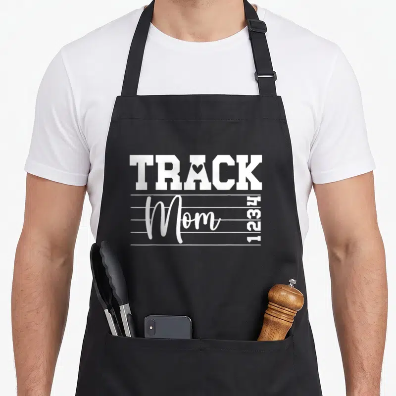 Field mother's apron - White TRACK Mom text with heart in A, four horizontal lines, and vertical 1234 numbers — shown on a black cotton canvas apron against a white background