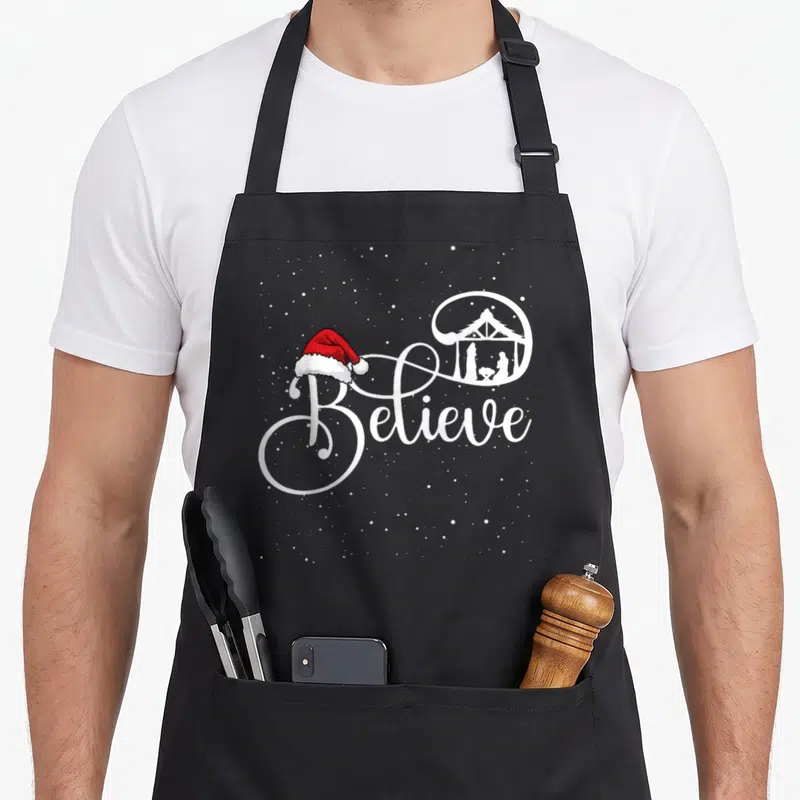 Believe family christmas aprons - Believe text with Santa hat and enclosed nativity scene surrounded by white snow dots — shown on a black cotton canvas apron against a white background