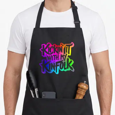 Kinfolk family reunion aprons - Stacked KICKIN IT WITH MY KINFOLK text in pink, yellow, green, blue, purple gradient — shown on a black cotton canvas apron against a white background