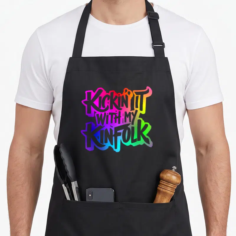 Kinfolk family reunion aprons - Stacked KICKIN IT WITH MY KINFOLK text in pink, yellow, green, blue, purple gradient — shown on a black cotton canvas apron against a white background
