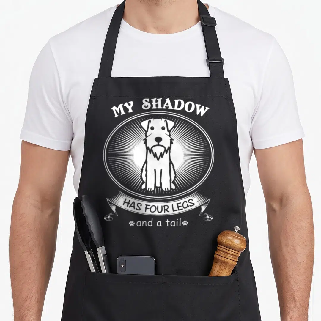 Dog miniature schnauzer apron - White line-art dog in oval frame with text MY SHADOW HAS FOUR LEGS and a tail — shown on a black cotton canvas apron against a white background