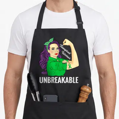 Warrior organ apron - Woman flexing arm with green ribbon and Transplant Warrior UNBREAKABLE text — shown on a black cotton canvas apron against a white background
