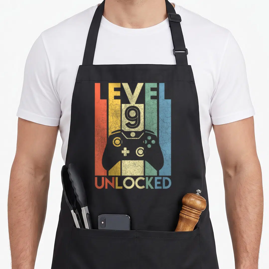 Video gamer 9th birthday apron - Retro game controller over red yellow green blue grey stripes with LEVEL 9 UNLOCKED — shown on a black cotton canvas apron against a white background