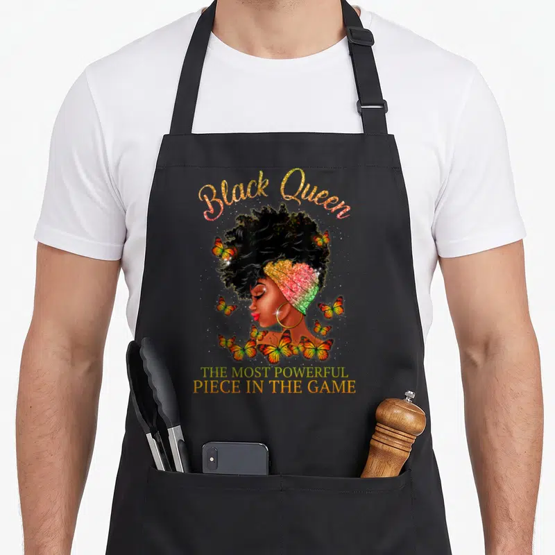 Most queen apron - Woman in rainbow headwrap surrounded by orange butterflies with gold Black Queen text — shown on a black cotton canvas apron against a white background