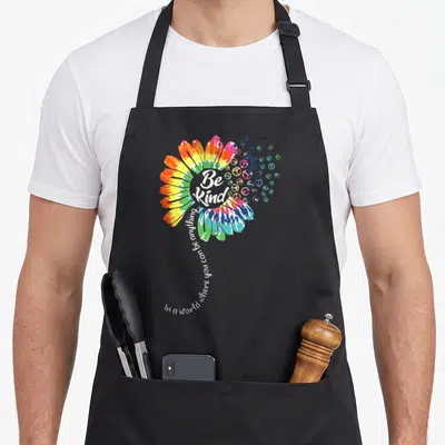 Unity bullying prevention apron - Tie-dye flower with Be Kind text and scattered peace signs in rainbow colors — shown on a black cotton canvas apron against a white background