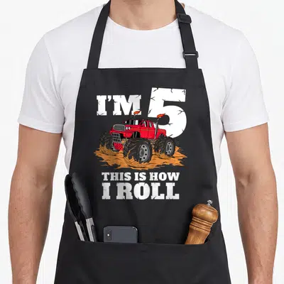 5th truck apron - Red monster truck over orange dirt with I'M 5 THIS IS HOW I ROLL text — shown on a black cotton canvas apron against a white background