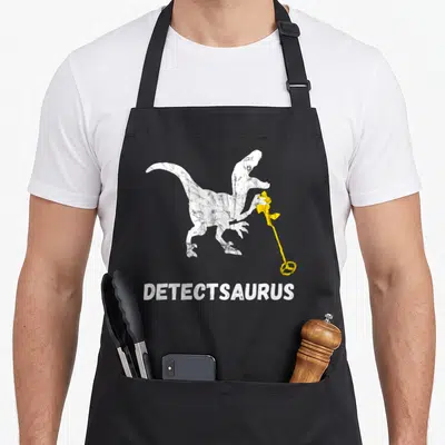 Saurus metal detecting apron - White distressed dinosaur holding yellow detector with DETECTSAURUS text below — shown on a black cotton canvas apron against a white background