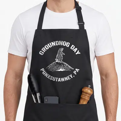 Hog groundhog apron - White groundhog on dirt mound with GROUNDHOG DAY PUNXSUTAWNEY PA distressed text — shown on a black cotton canvas apron against a white background