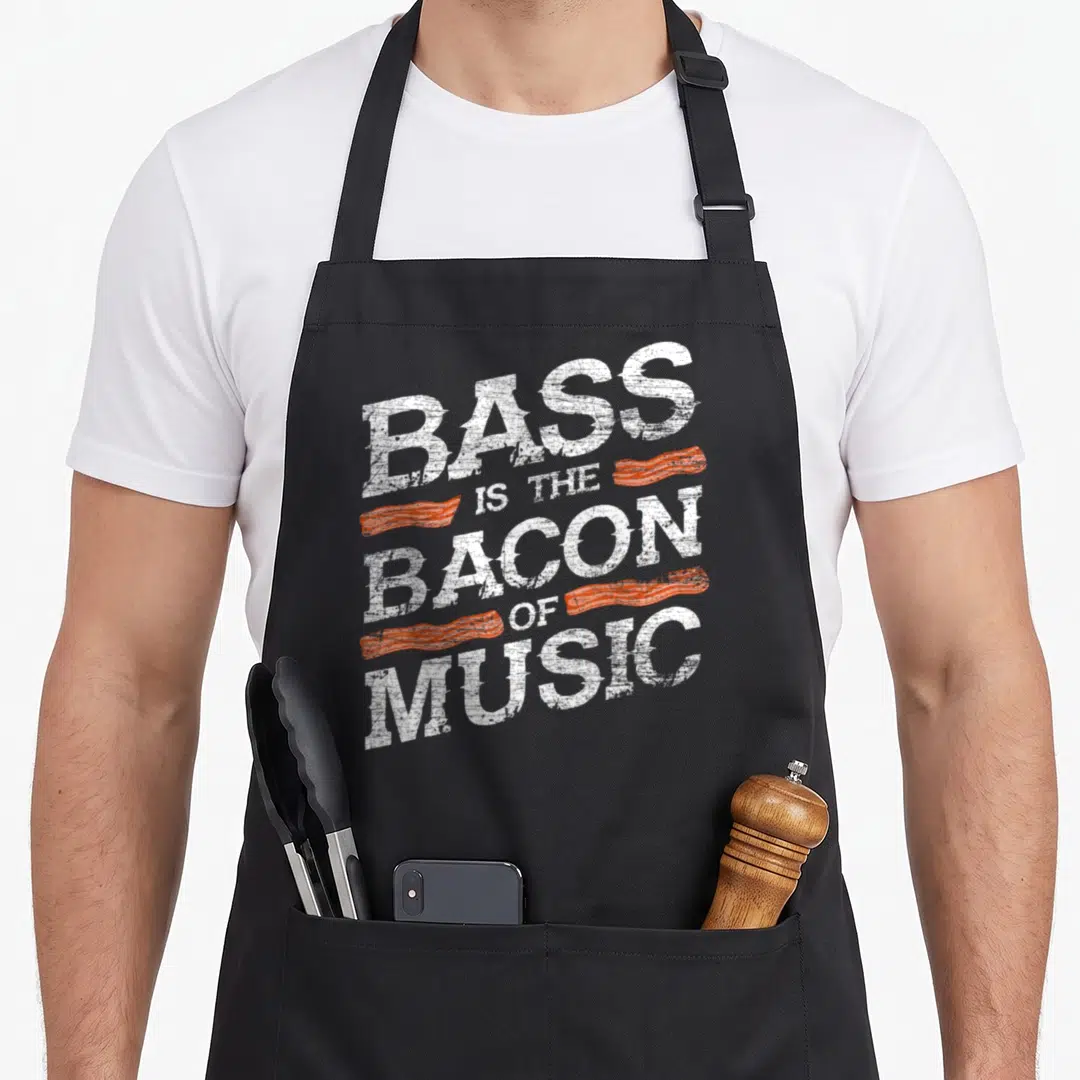 Music bacon apron - Distressed white text reading Bass Is The Bacon Of Music with four orange bacon strips — shown on a black cotton canvas apron against a white background