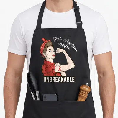 Warrior aneurysm awareness apron - Cartoon woman flexing arm with red ribbon and text Brain Aneurysm warrior UNBREAKABLE — shown on a black cotton canvas apron against a white background