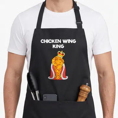 Fan chicken apron - Cartoon fried chicken wing wearing a red cape and gold crown under white text CHICKEN WING KING — shown on a black cotton canvas apron against a white background