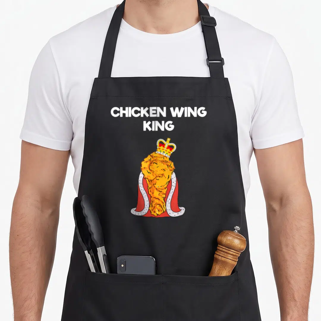 Fan chicken apron - Cartoon fried chicken wing wearing a red cape and gold crown under white text CHICKEN WING KING — shown on a black cotton canvas apron against a white background