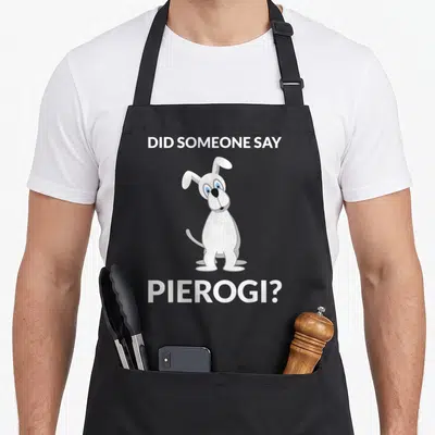 Pierogi polish kitchen aprons - White cartoon dog between DID SOMEONE SAY PIEROGI text on grey — shown on a black cotton canvas apron against a white background