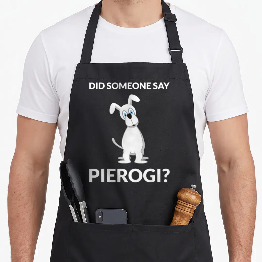 Pierogi polish kitchen aprons - White cartoon dog between DID SOMEONE SAY PIEROGI text on grey — shown on a black cotton canvas apron against a white background