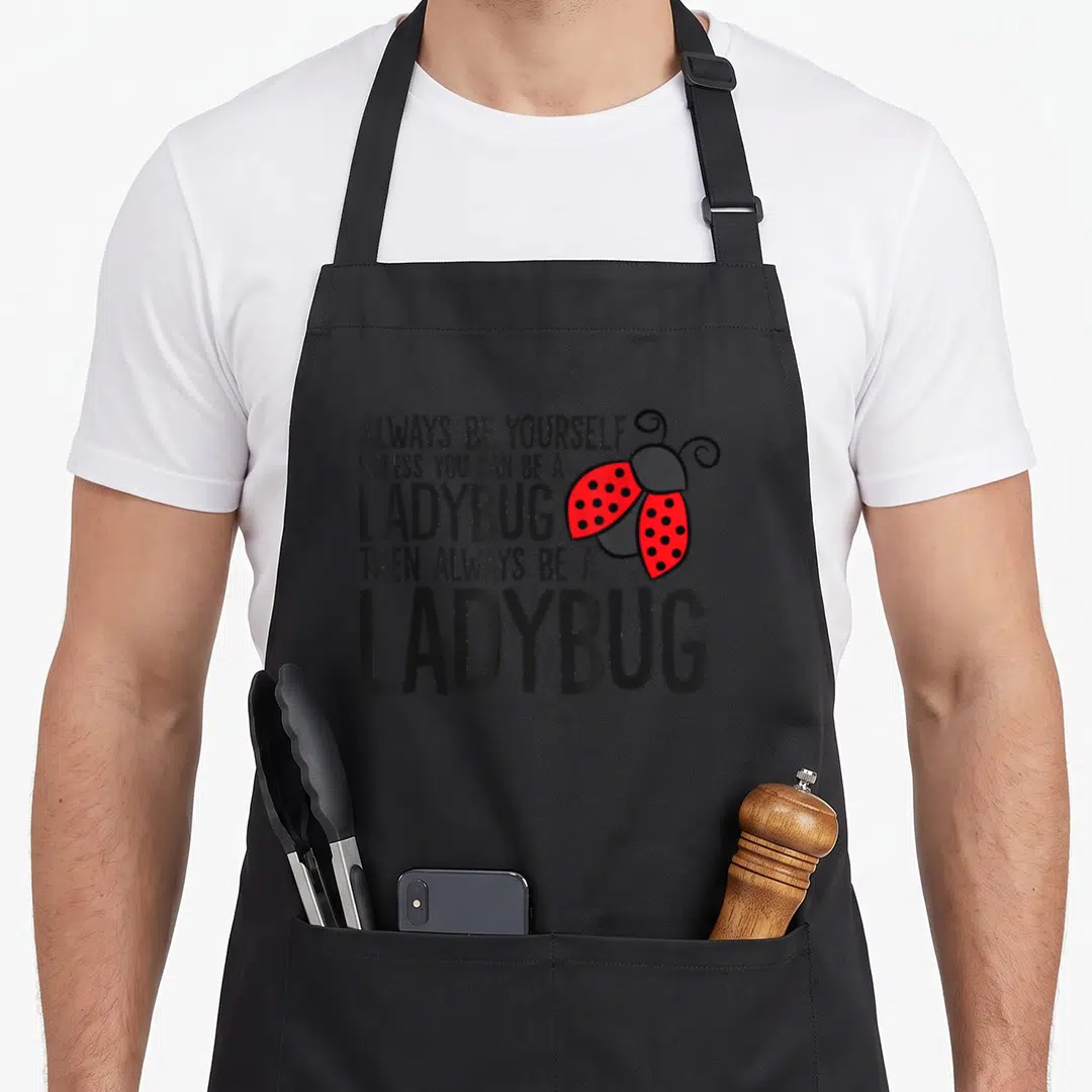 Yourself ladybug apron - Cartoon ladybug with red spotted wings next to black text saying ALWAYS BE YOURSELF UNLESS YOU — shown on a black cotton canvas apron against a white background