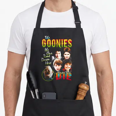 Edit character aprons - The Goonies title, four boy faces, One Eyed Willy portrait, and movie quotes in yellow red blue — shown on a black cotton canvas apron against a white background