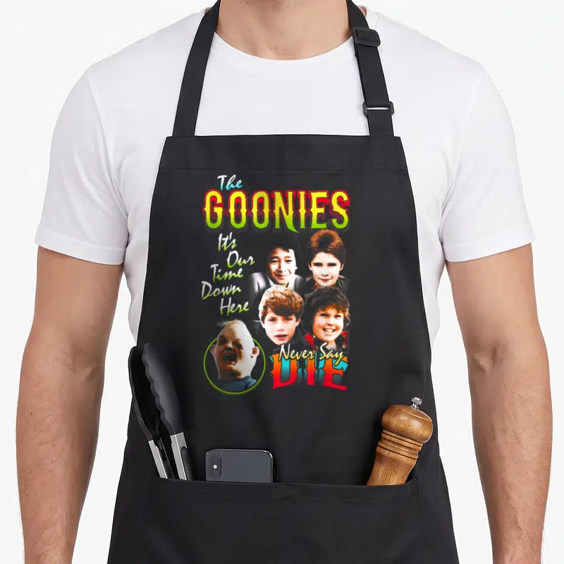 Edit character aprons - The Goonies title, four boy faces, One Eyed Willy portrait, and movie quotes in yellow red blue — shown on a black cotton canvas apron against a white background