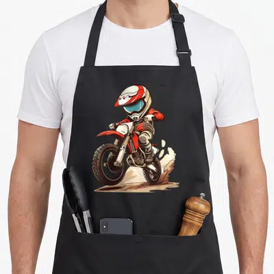 Vintage dirt bike apron - Cartoon motocross rider in red and white gear kicking up brown dirt dust cloud — shown on a black cotton canvas apron against a white background
