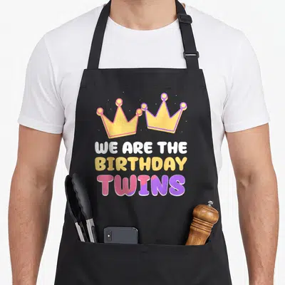Twin celebrate birthday apron - Two cartoon crowns above stacked text WE ARE THE BIRTHDAY TWINS in yellow pink purple — shown on a black cotton canvas apron against a white background
