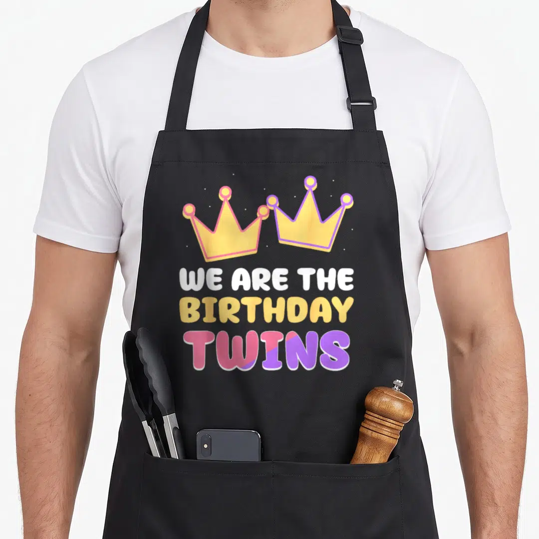 Twin celebrate birthday apron - Two cartoon crowns above stacked text WE ARE THE BIRTHDAY TWINS in yellow pink purple — shown on a black cotton canvas apron against a white background