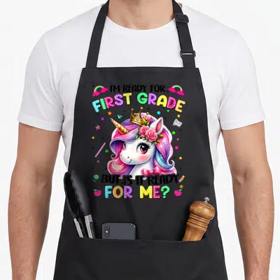 Grade school unicorn apron - Cartoon unicorn with rainbow mane surrounded by school supplies and polka-dot text reading — shown on a black cotton canvas apron against a white background