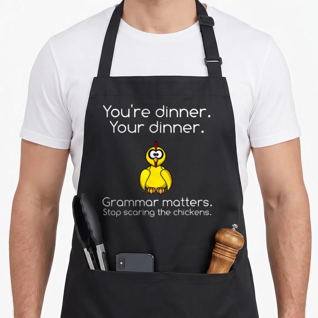 Grammar chicken apron - Cartoon yellow chicken between You're dinner Your dinner and Stop scaring the chickens text — shown on a black cotton canvas apron against a white background