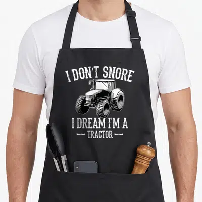 Dream tractor apron - Distressed white text I DON'T SNORE I DREAM I'M A TRACTOR surrounding a black tractor illustration — shown on a black cotton canvas apron against a white background