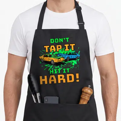 Hit auto apron - Don't Tap It Hit It Hard text surrounding two crashing orange and blue cars on green splatter — shown on a black cotton canvas apron against a white background